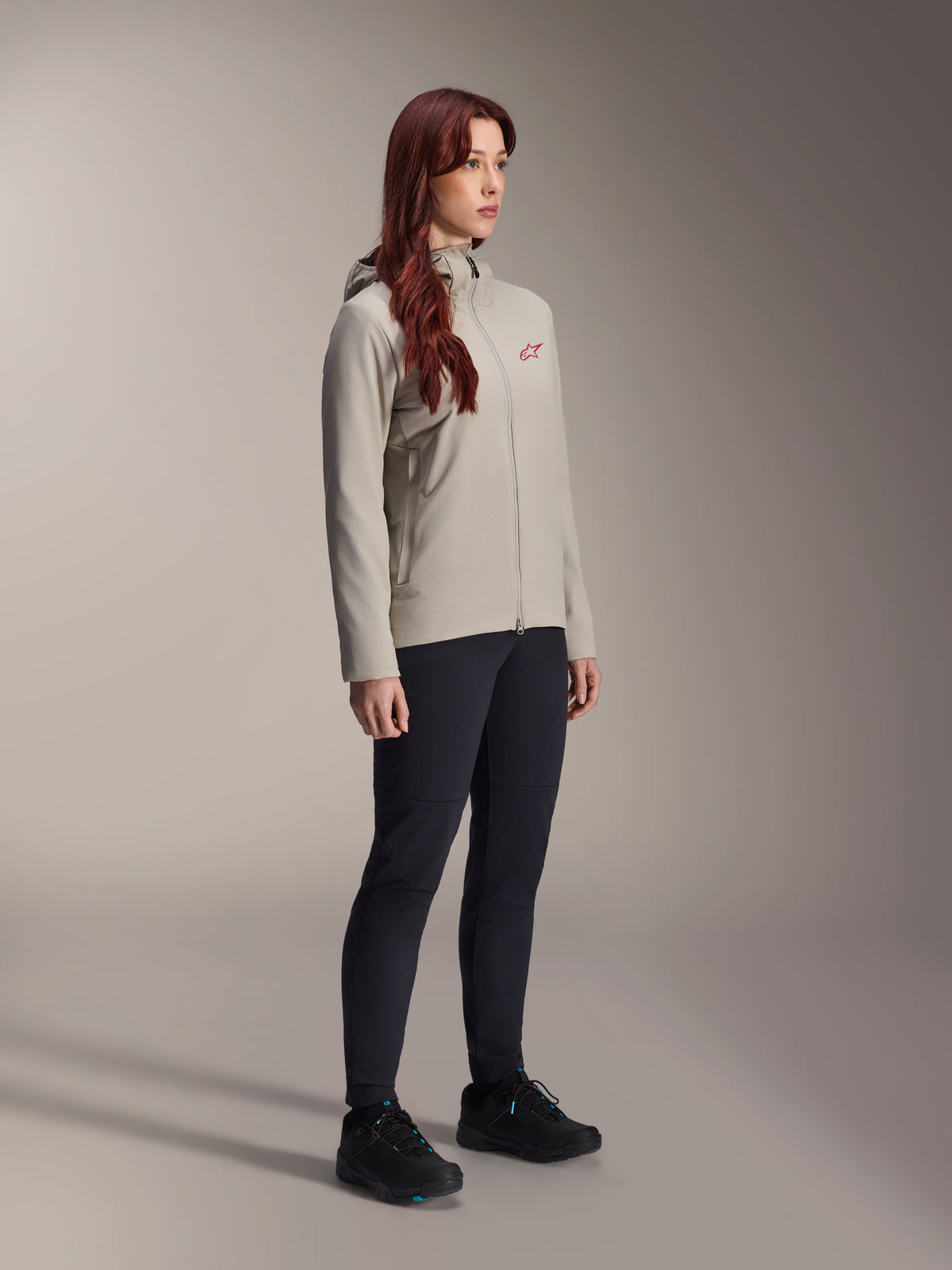 Alpinestars Stella A-Dura Thermal Jacket, titaniumgrijs, thermische fietsjas voor dames met een rood sterlogo op de borst, gecombineerd met een zwarte technische broek en zwarte mountainbikeschoenen, studio-opname voor outdoor-fietskleding