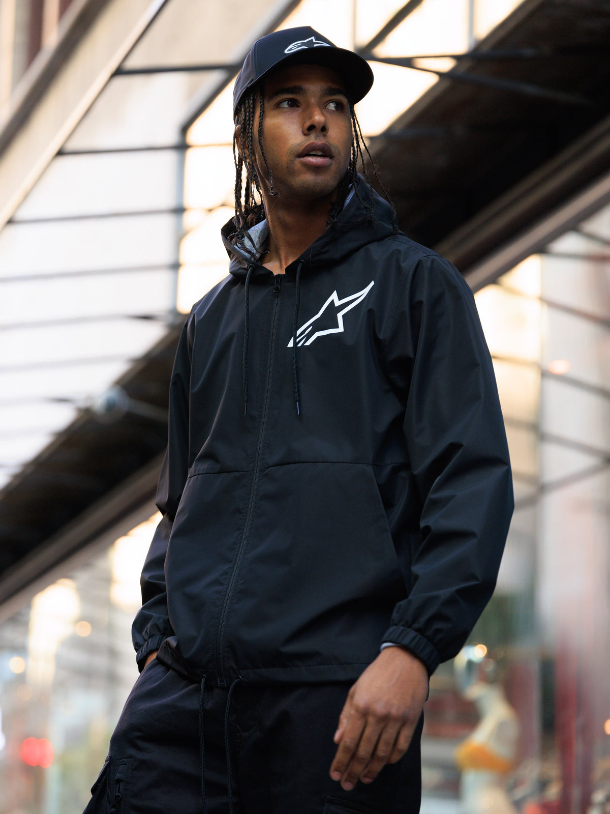 Alpinestars Speeder Chromium Windbreak Jacket, zwart, windbreaker met capuchon en een wit logo op de borst, gedragen door een man met een bijpassende zwarte Alpinestars pet in een stedelijke omgeving, casual motorsport streetwear.