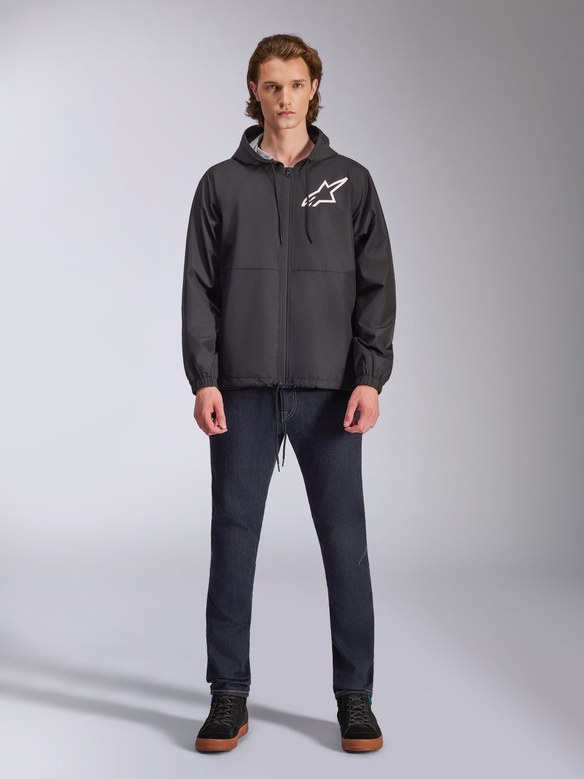 Alpinestars Speeder Chromium Windbreak Jacket, zwarte windbreaker met wit Astars-logo, gedragen door een model met donkerblauwe jeans en zwarte sneakers, voorzien van een capuchon en ritssluiting aan de voorzijde, casual motorsport-geïnspireerde streetwear