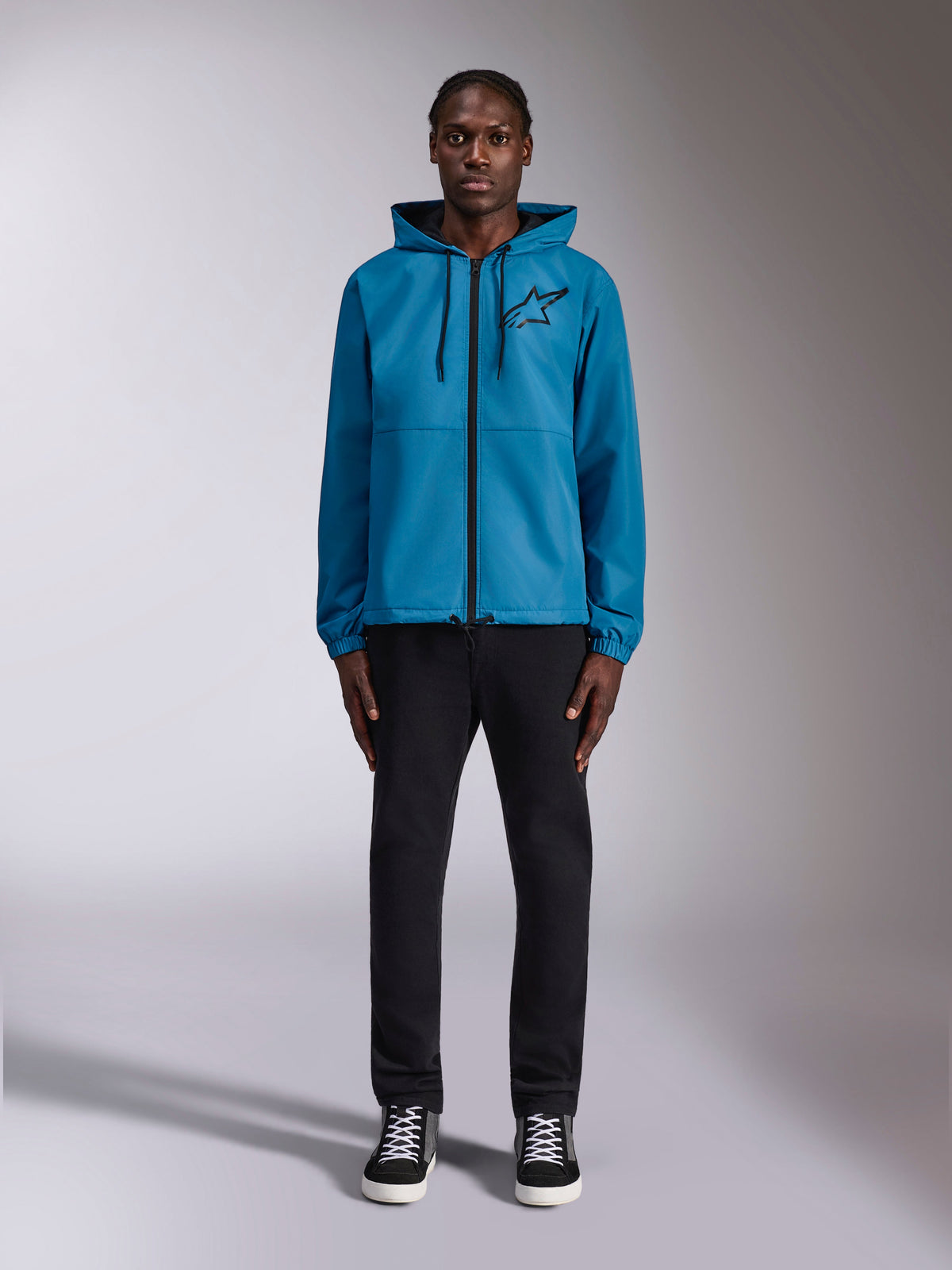 Model wearing the Alpinestars Speeder Chromium Windbreak Jacket in Blue, featuring a hooded design with black drawcords, a black front zipper, and a black Astars chest logo, paired with black pants and sneakers, casual motorsport streetwear
