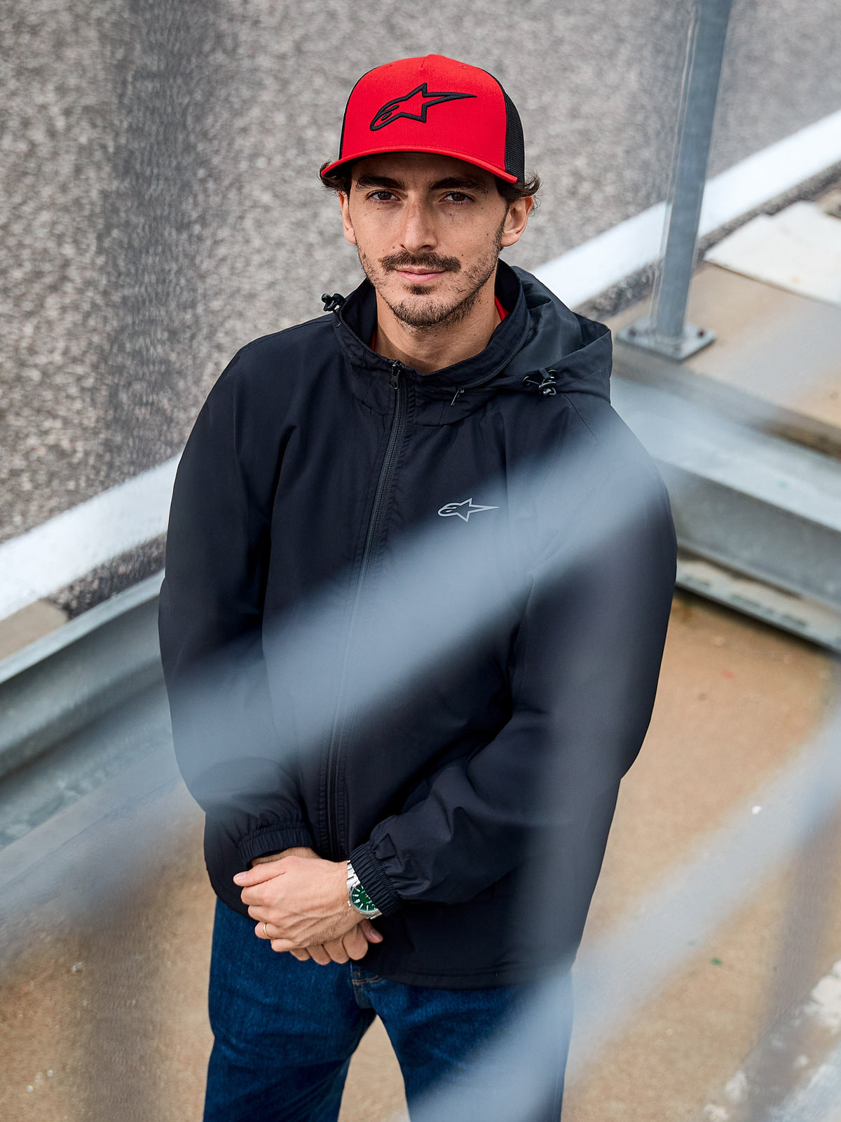 Man die de Alpinestars Overhaul Racer Jacket in het zwart draagt, een waterbestendig regenjack met capuchon en een klein logo op de borst, gecombineerd met een rode Alpinestars pet, casual motorsport lifestyle kleding