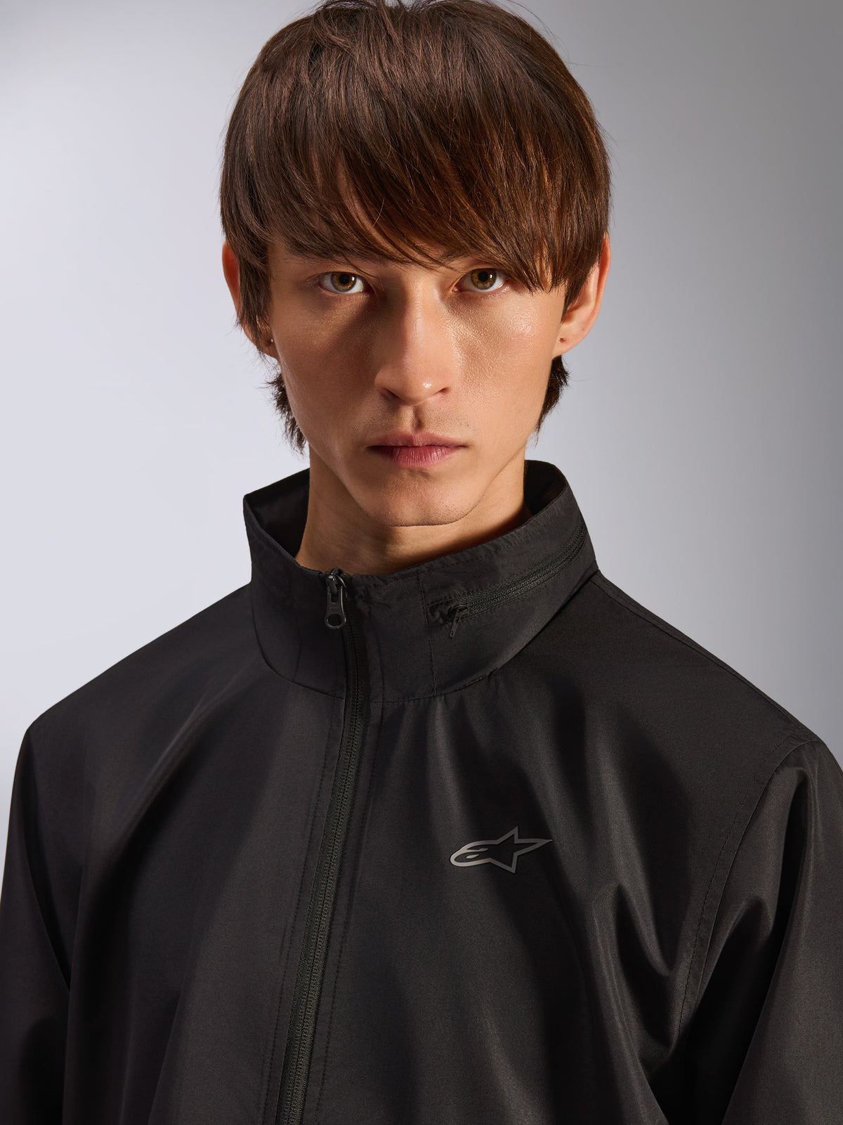 Alpinestars Overhaul Racer Jacket, zwarte regenjas, close-up portret van een mannelijk model dat de jas draagt, met een hoge kraag en een klein zilveren Astars-logo op de borst, motorsport-geïnspireerde casual kleding