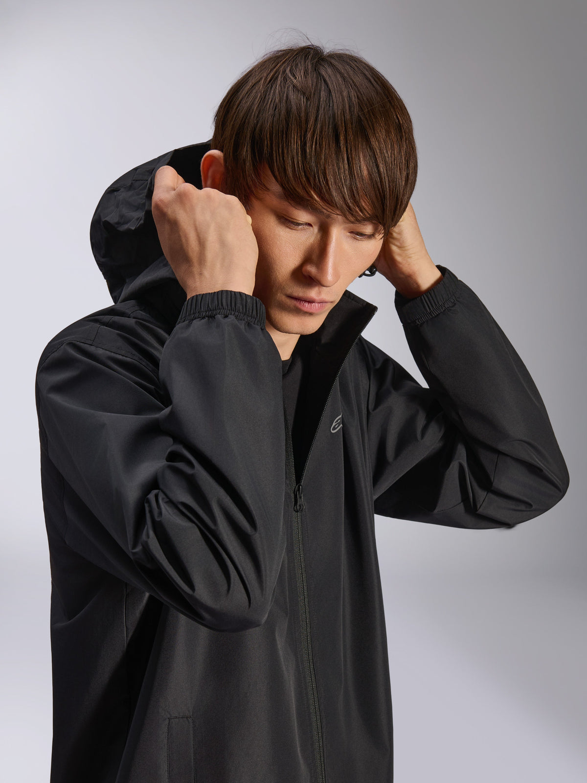 Alpinestars Overhaul Racer Jacket, zwarte regenjas met capuchon, gedragen door model met de capuchon op, voorzien van waterbestendige 2.5L stof, elastische manchetten en een subtiel logo op de borst, casual motorsport-geïnspireerde buitenkleding