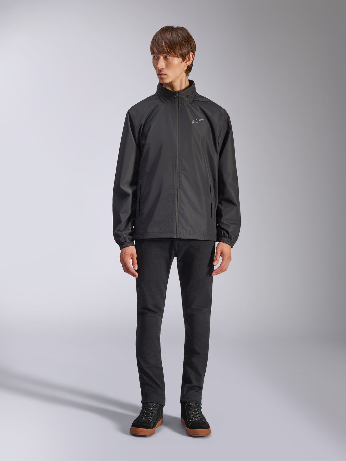 Alpinestars Overhaul Racer Jacket, zwarte regenjas voor heren, volledige weergave op een model gecombineerd met een zwarte broek en casual sneakers, voorzien van een klein wit logo op de borst, waterbestendige stof en een casual op motorsport geïnspireerd ontwerp