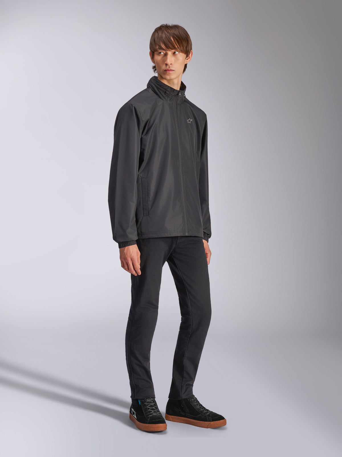 Alpinestars Overhaul Racer Jacket, zwart regenjack, gedragen met een zwarte broek en zwarte sneakers met gumzolen, voorzien van een wit ster-logo op de borst, ritssluiting aan de voorzijde, casual op motorsport geïnspireerde sportkleding