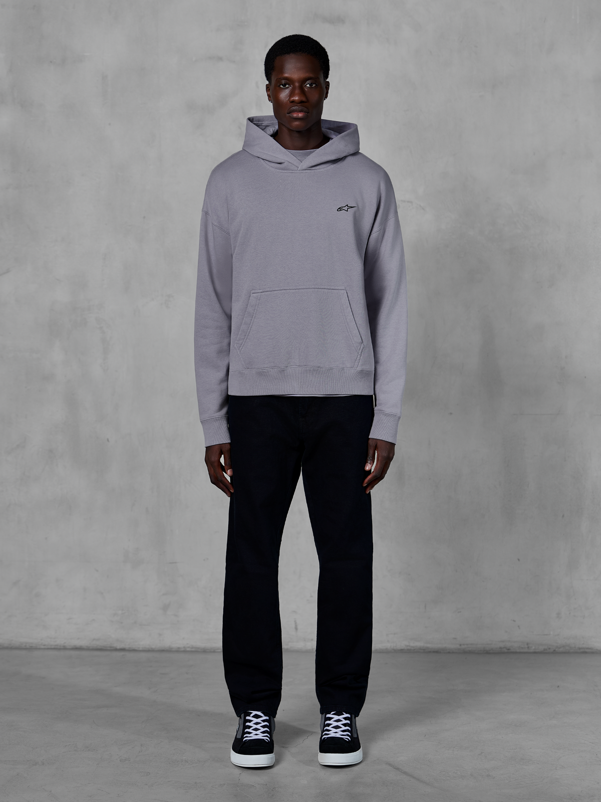 Alpinestars Essentials Ageless Hoodie, zilver, mannelijk model in casual streetwear, klein zwart Alpinestars-logo op de borst, kangoeroezak aan de voorzijde, minimalistisch ontwerp, gecombineerd met zwarte broek en sneakers.