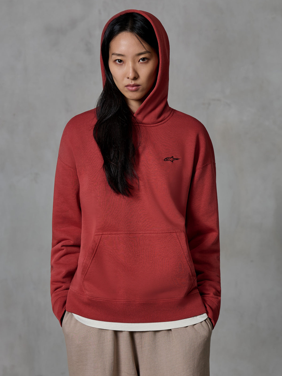 Alpinestars Essentials Ageless Hoodie, baksteenrode pullover sweatshirt, gedragen door een vrouwelijk model met de capuchon op, klein zwart geborduurd sterlogo op de borst, kangoeroezak, casual lifestyle streetwear geïnspireerd op de motorsport