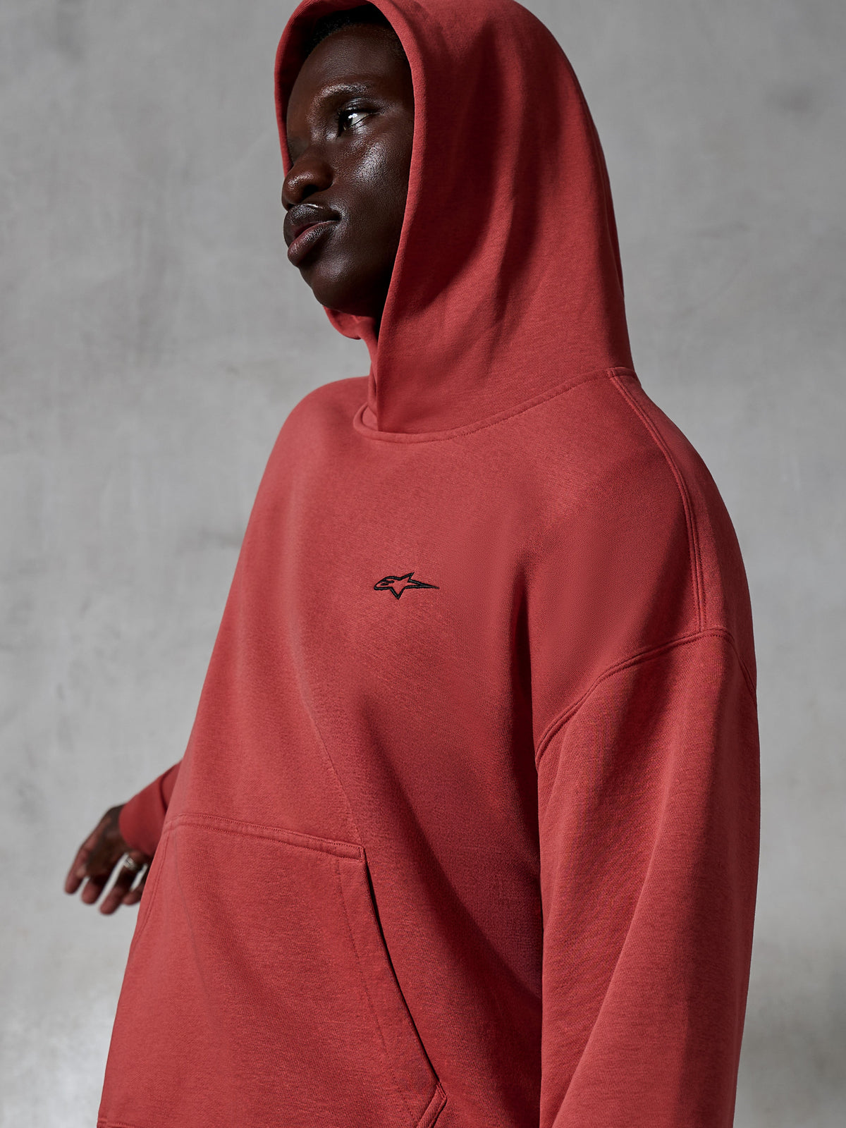 Alpinestars Essentials Ageless Hoodie, casual sportkleding, steenrood, gedragen door een model met de capuchon op, voorzien van een klein zwart geborduurd logo, zachte fleece textuur, relaxed fit streetwear