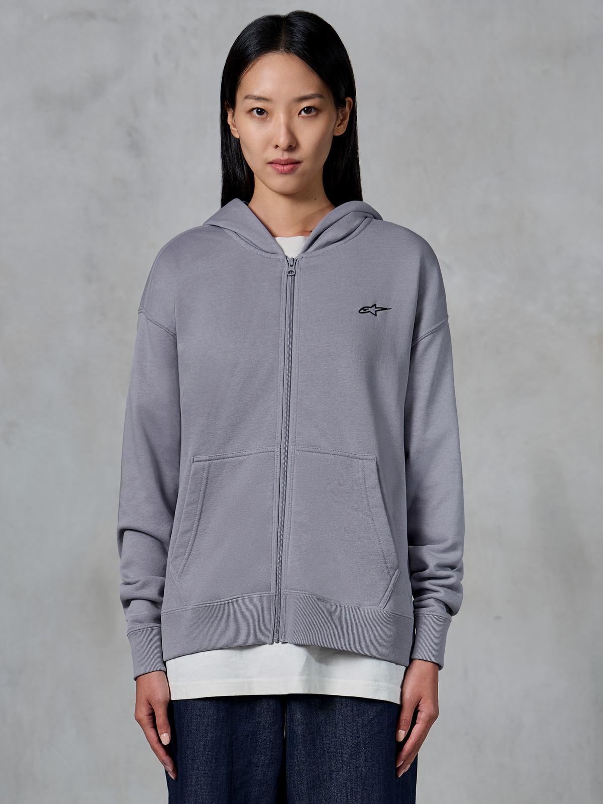 Alpinestars Essentials Ageless Zip Hoodie, Silver, casual streetwear, fleece sweatshirt met volledige ritssluiting en een klein zwart Alpinestars-logo op de borst, gedeelde kangoeroezak, gedragen door model over een wit t-shirt en donkere jeans