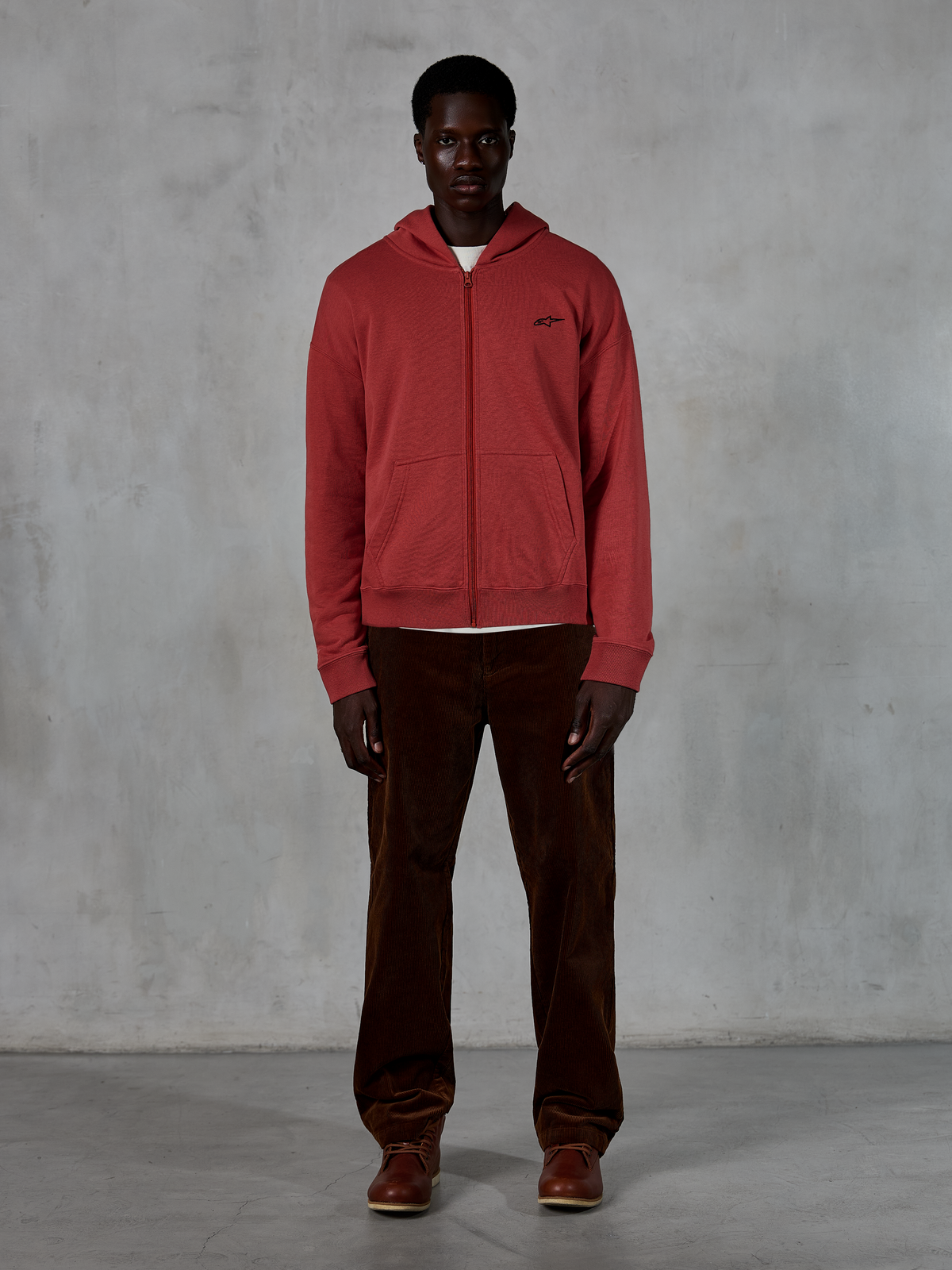 Alpinestars Essentials Ageless Zip Hoodie, casual streetwear, brick red with small black star logo, full-zip front, worn by a model with brown corduroy pants and boots, premium motorsport-inspired design