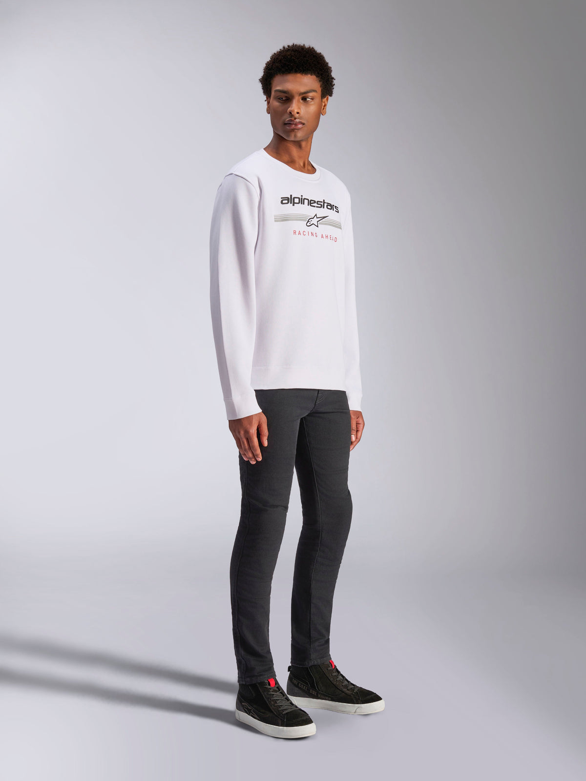 Alpinestars Bettern Crew, witte pullover sweatshirt, met zwart Alpinestars-logo en rode 'Racing Ahead'-tekst, casual motorsport streetwear, gedragen door een model met donkere broek en zwarte sneakers