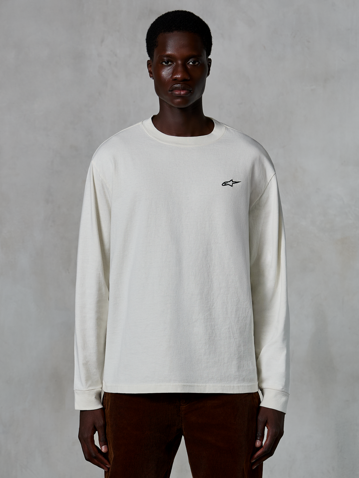 Alpinestars Essentials Ageless LS Tee, T-shirt met lange mouwen, gebroken wit, klein zwart sterlogo op de borst, casual streetwear, ontspannen pasvorm, gedragen door een model