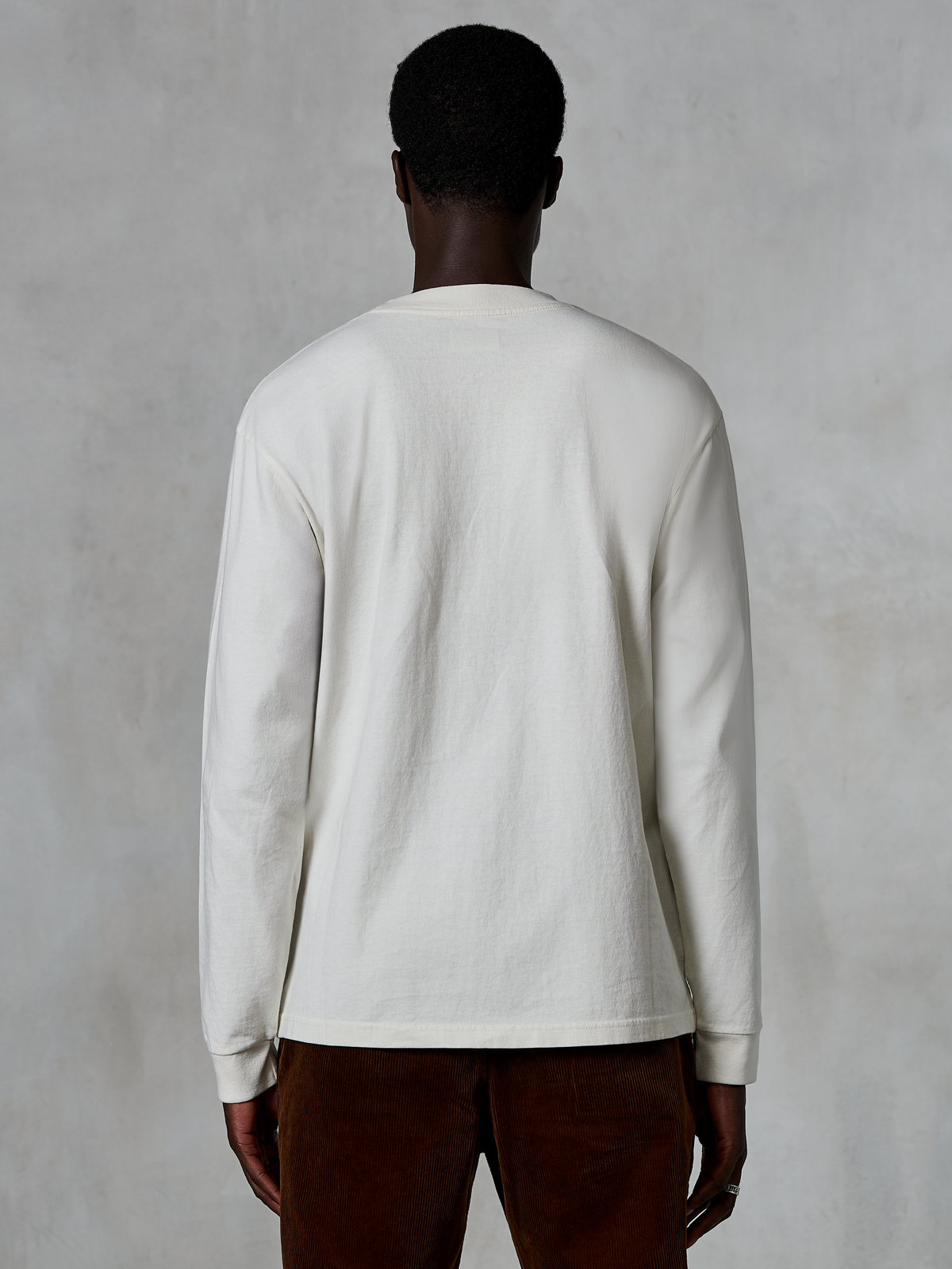 Alpinestars Essentials Ageless LS Tee, T-shirt met lange mouwen, Off White, achteraanzicht van een persoon die het shirt draagt met een bruine corduroy broek, ontspannen pasvorm, getextureerde biologische katoenen stof, minimalistische casual streetwear