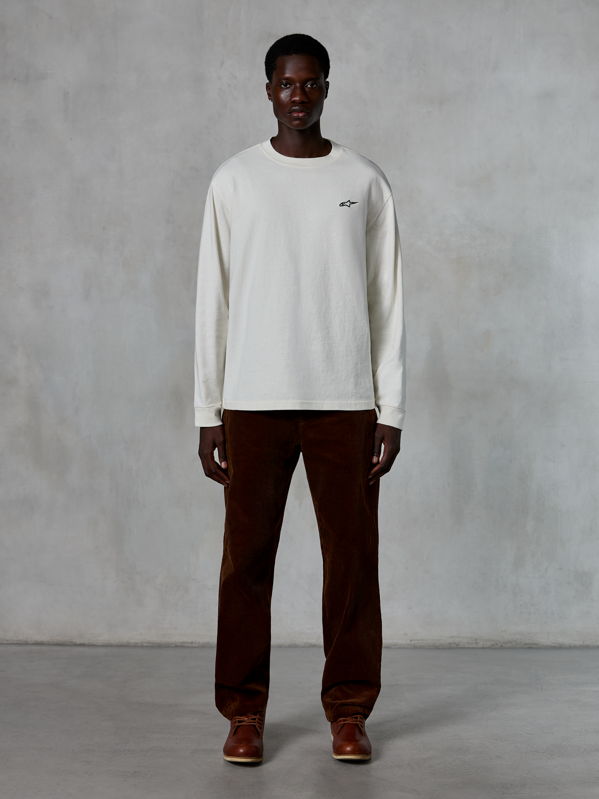 Alpinestars Essentials Ageless LS Tee, T-shirt met lange mouwen, Off-White, gedragen door model met bruine corduroy broek en bruine leren laarzen, met een klein zwart logo op de borst, ontspannen eigentijdse pasvorm, casual lifestyle streetwear