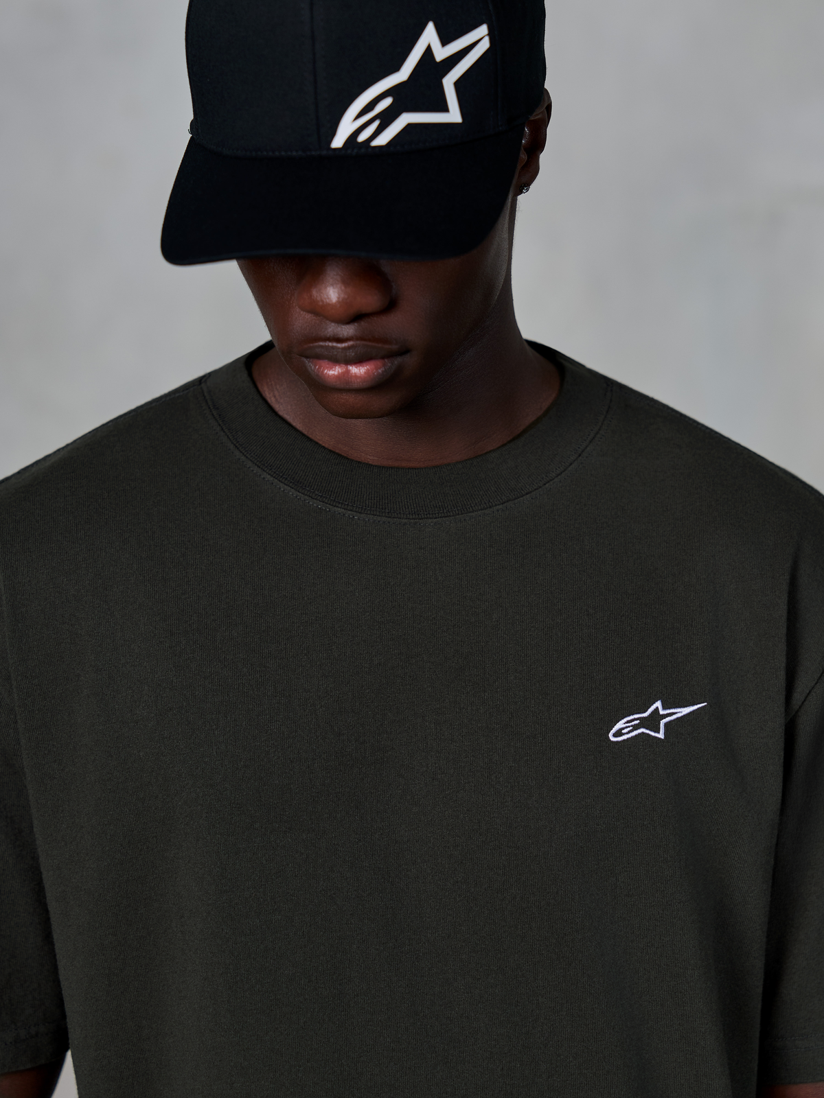 Alpinestars Essentials Ageless SS Tee, Short Sleeve T-Shirt, Off Black, featuring a small white Alpinestars logo on the chest, worn with a black logo baseball cap, casual motorsport streetwear style