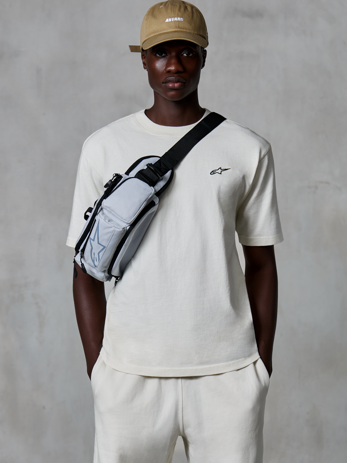 Alpinestars Essentials Ageless SS Tee, T-shirt met korte mouwen, gebroken wit, gedragen met bijpassende shorts en een beige pet, voorzien van een klein zwart logo op de borst en een grijze crossbodytas, casual streetwear geïnspireerd op de motorsport