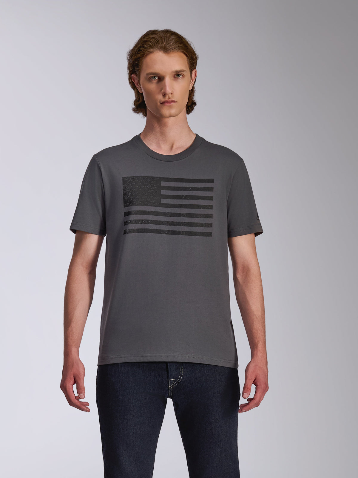 Alpinestars Waving CSF Tee, T-shirt met korte mouwen, charcoal, met een zwarte op een vlag geïnspireerde Blaze-logo graphic op de borst, gedragen door een model met een donkere jeans, casual motorsport streetwear