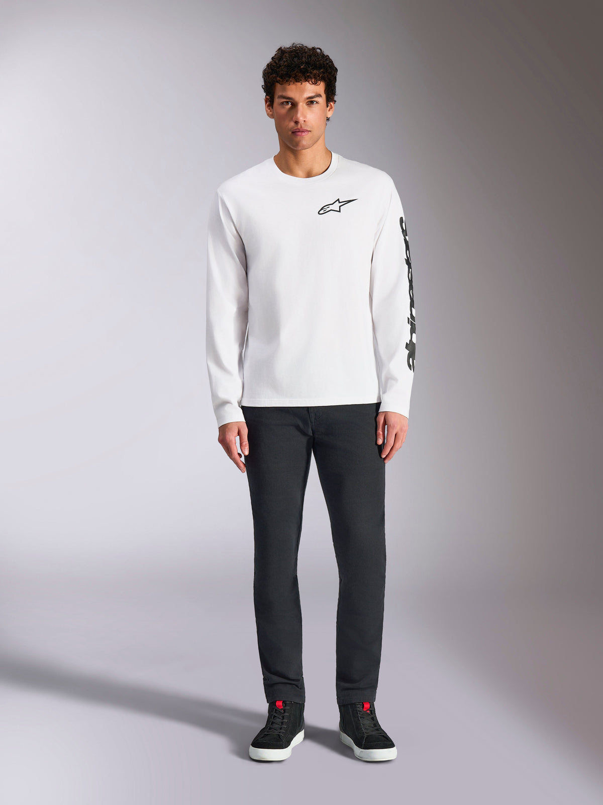 Alpinestars Trackside CSF Tee - lange mouwen, wit met zwarte branding, klein logo op de borst en verticale tekst op de mouw, gedragen door model met donkere broek en zwarte sneakers, casual op motorsport geïnspireerde streetwear