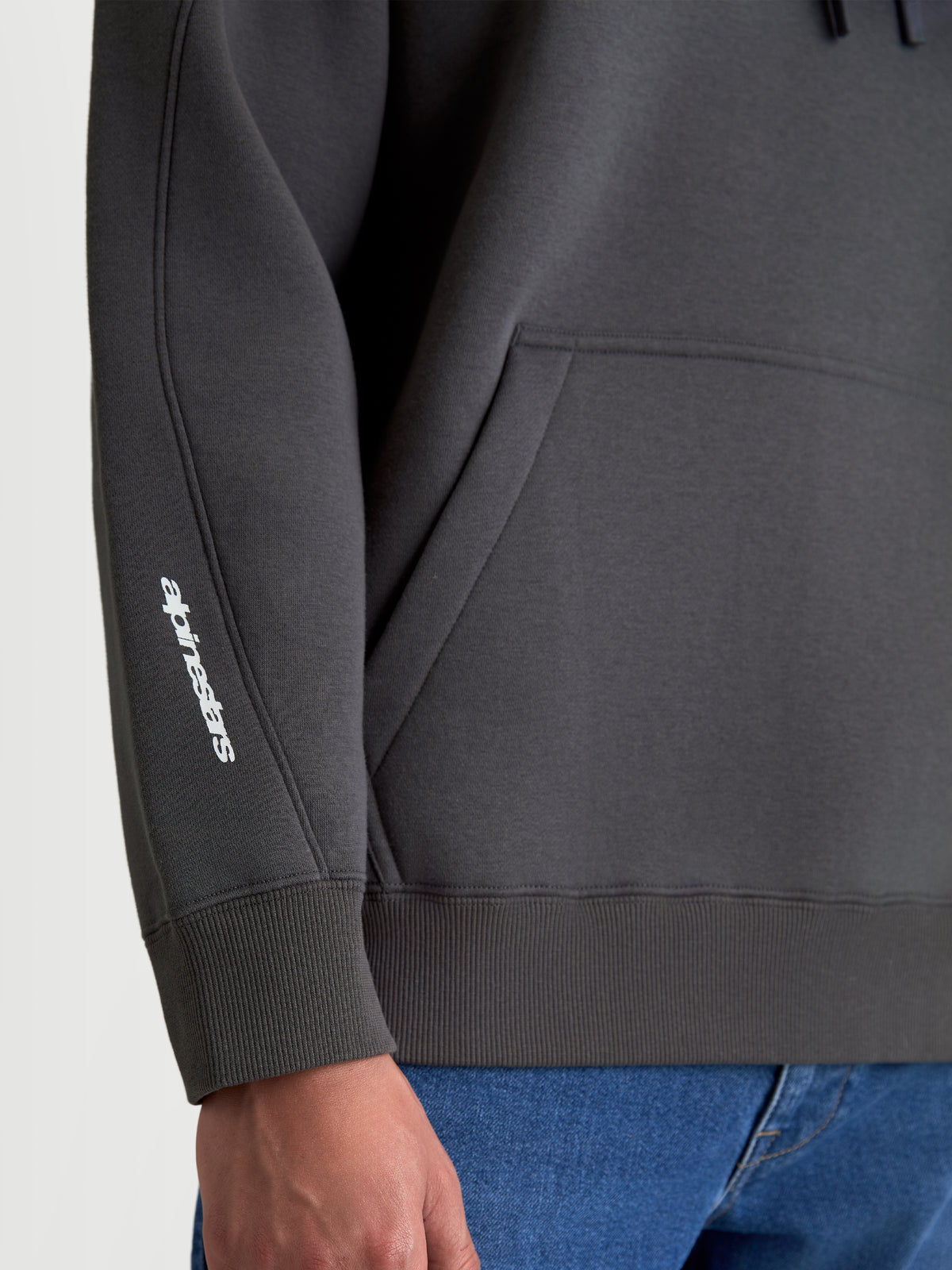 Alpinestars Prelude Oversized Hoodie, Sportswear Fleece Pullover in Charcoal Gray, detail shot showing the white puff-print logo on the sleeve, decorative seaming, and ribbed cuff and hem.