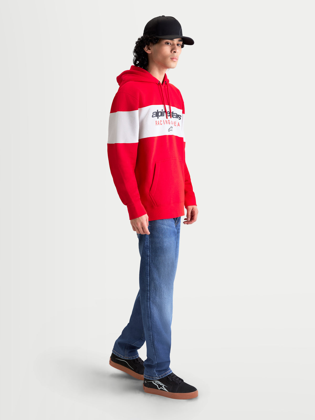 Alpinestars Cubic Hoodie, heren-sportswear fleece pullover in rood en wit, met kleurgeblokt design en Alpinestars woordlogo op de borst. Zij-aanzicht op model met capuchon met trekkoord, kangoeroezak en geribde manchetten voor casual comfort naast de motor.