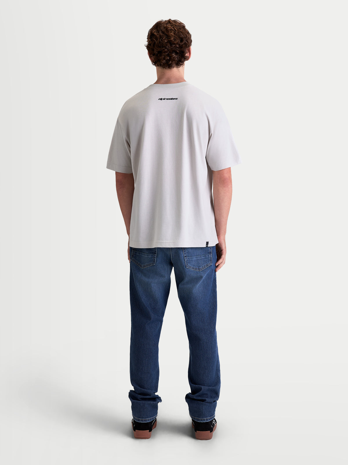 Alpinestars Prelude Oversized CSF T-shirt, heren-sportswear t-shirt met korte mouwen, cementgrijs, achteraanzicht op model met klein zwart Alpinestars-logo en een relaxte oversized pasvorm, hoogwaardige katoen-polyestermix voor een casual look naast de motor.