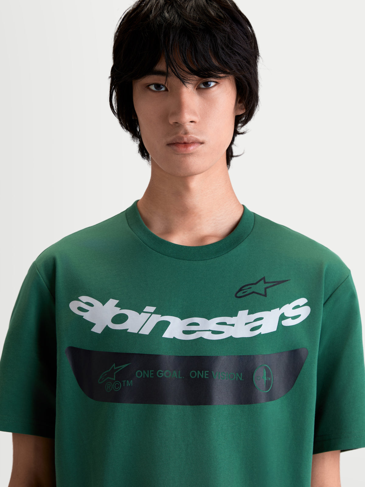 Alpinestars Singular CSF T-shirt met korte mouwen, heren t-shirt op model in groen, voorzien van een wit gebogen Alpinestars woordmerk en een zwarte band met het One Goal One Vision motto, premium katoenmix casual sportswear.