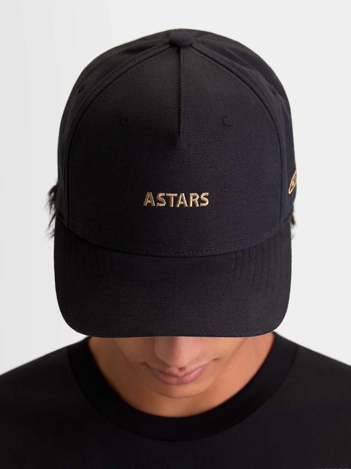 Alpinestars County Canvas pet, zwarte sportswearpet met goudkleurig geborduurd ASTARS-logo, gebogen klep en gestructureerde kroon, unisex motorsport-geïnspireerde vrijetijdspet, oxford peached stof, vooraanzicht op model.