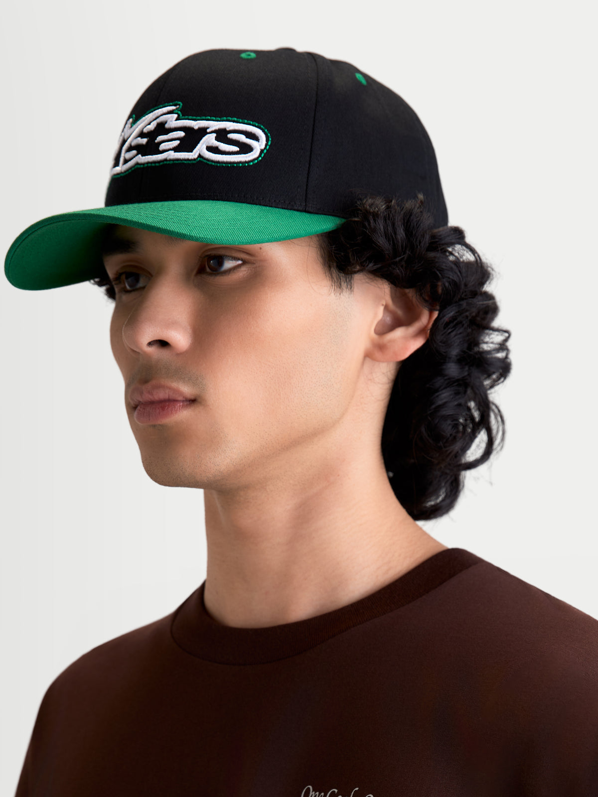Alpinestars Retro Snapback pet, sportswear pet gedragen door herenmodel, zwart en groen, met gebogen klep, gestructureerde kroon en geborduurd Astars-logo met groen stiksel, premium katoenen keperstof.