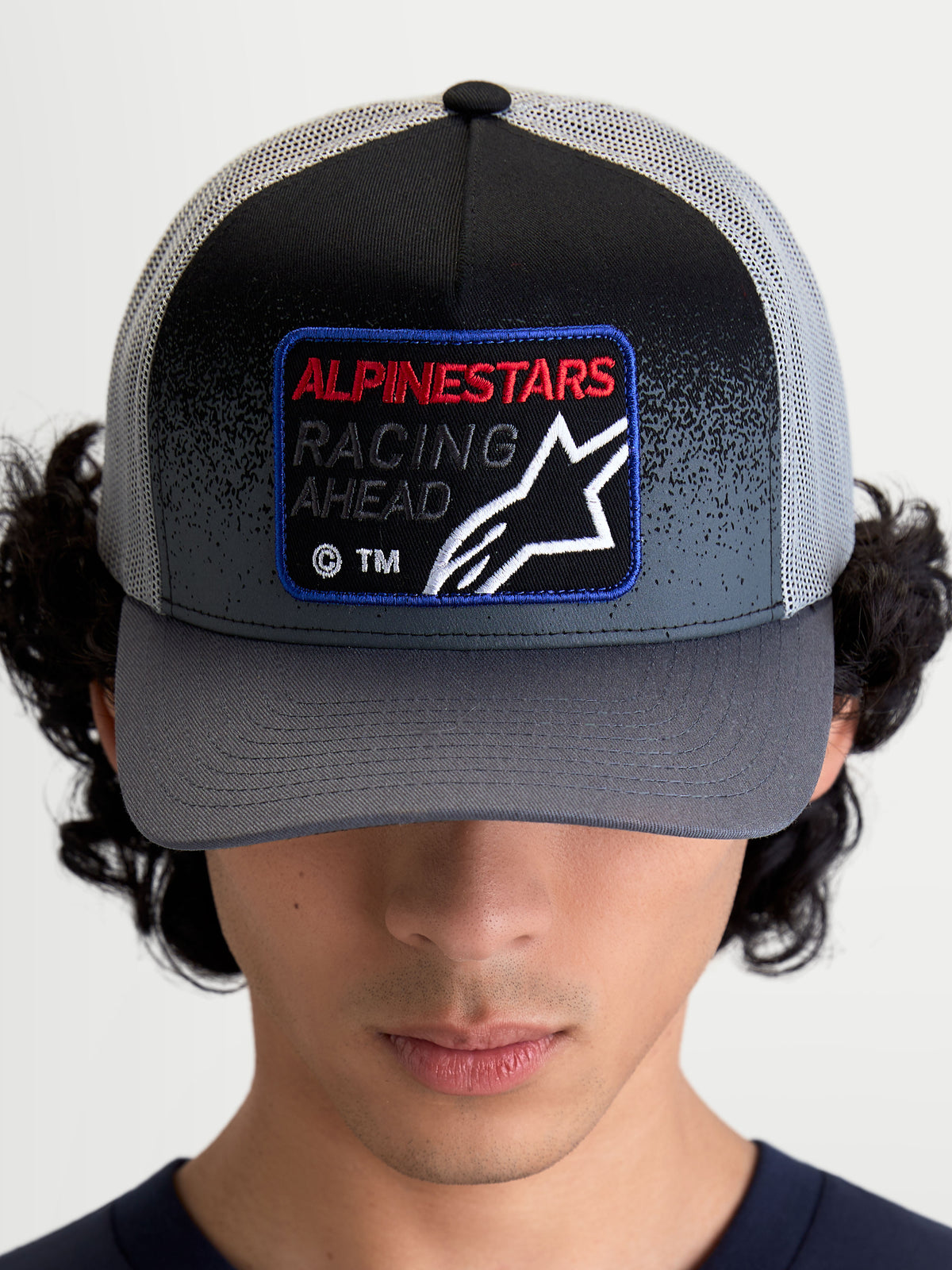 Alpinestars Gravel Trucker pet, sportswear trucker pet, donkergrijs en zwart, met gebogen klep en geweven patch op de voorzijde met 'Racing Ahead' en Astars-logo, vooraanzicht op model.