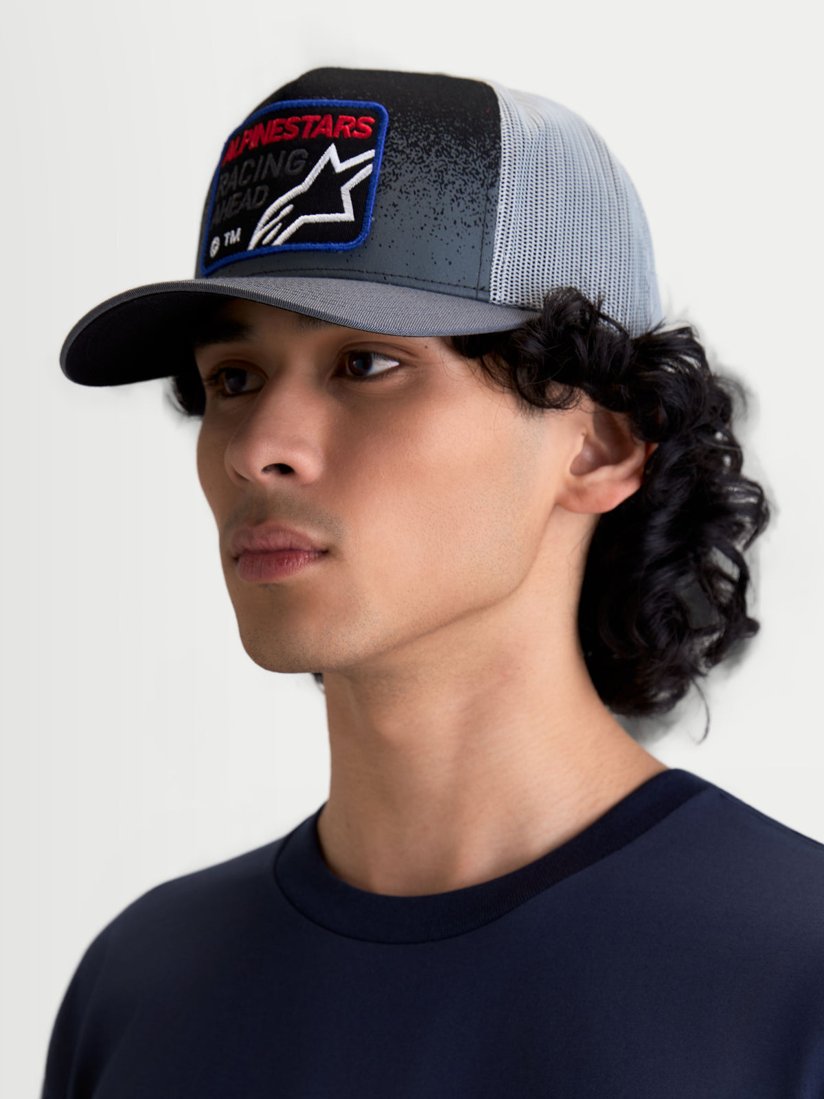 Alpinestars Gravel Trucker pet, casual sportswear pet op model, donkergrijs en zwart met lichtgrijze mesh achterkant, gestructureerde kroon, gebogen klep en geweven frontpatch met rode Alpinestars-tekst, motorsports-geïnspireerde stijl.
