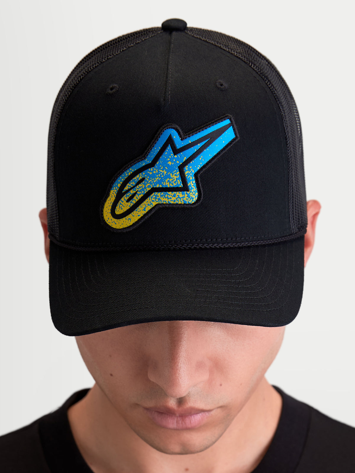 Alpinestars Meteorite Trucker pet, sportswear trucker pet met een gestructureerde kroon en gebogen klep, zwart met een blauwe Astars-logo patch met grindtextuur, gedragen door een model.