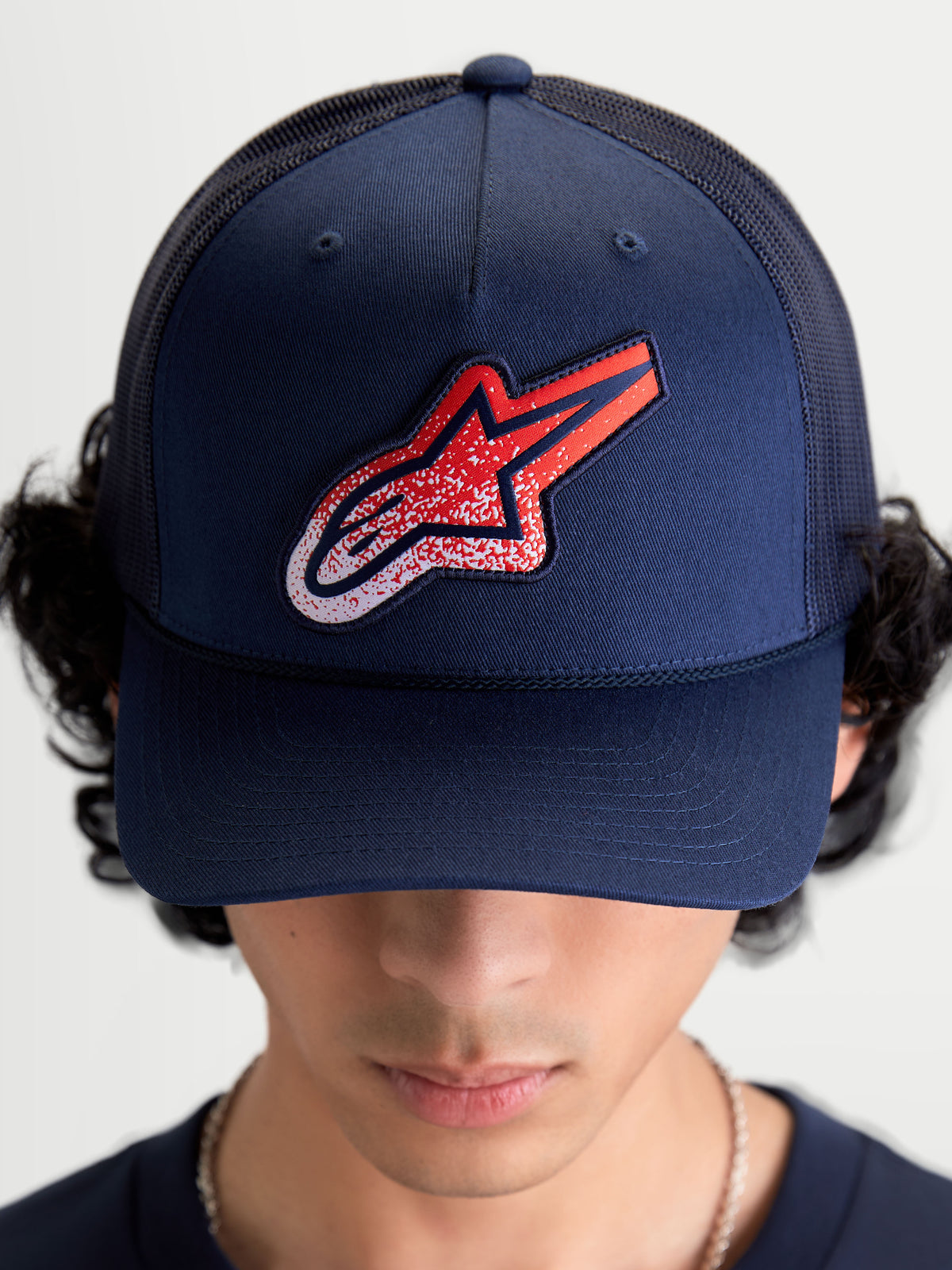 Alpinestars Meteorite Trucker pet, sportieve mesh pet voor heren en dames, marineblauw met rood en wit Astars-logo met grindtextuur, gestructureerde kroon en gebogen klep, vooraanzicht op model.