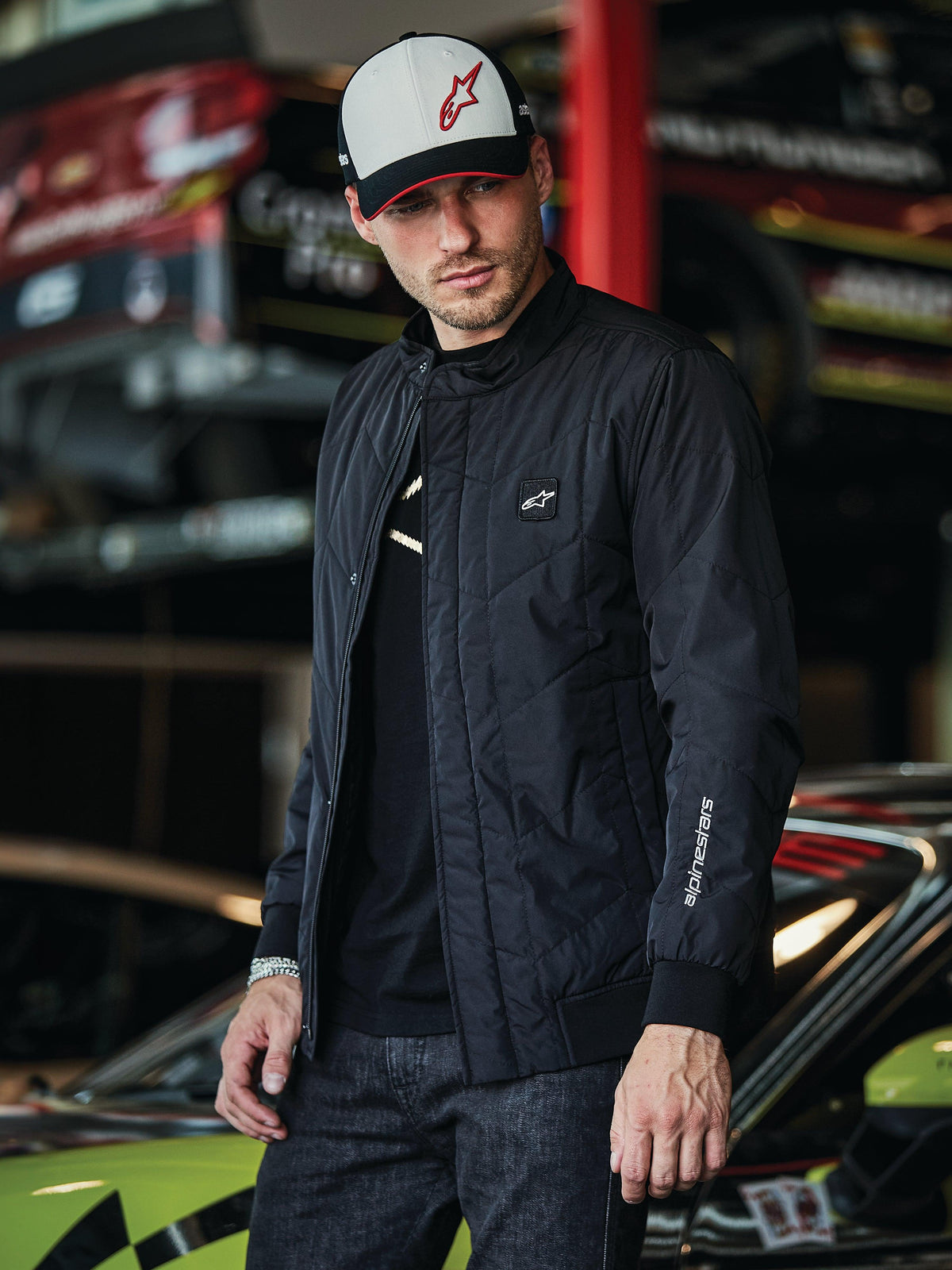 Alpinestars Precedent jas, zwarte gesteppte jas met Formule 1-styling, voorzien van een iconisch schuin gestikt patroon, logopatch op de borst en wit borduurwerk op de mouw, gedragen met een bijpassende Alpinestars pet in een motorsport garageomgeving.
