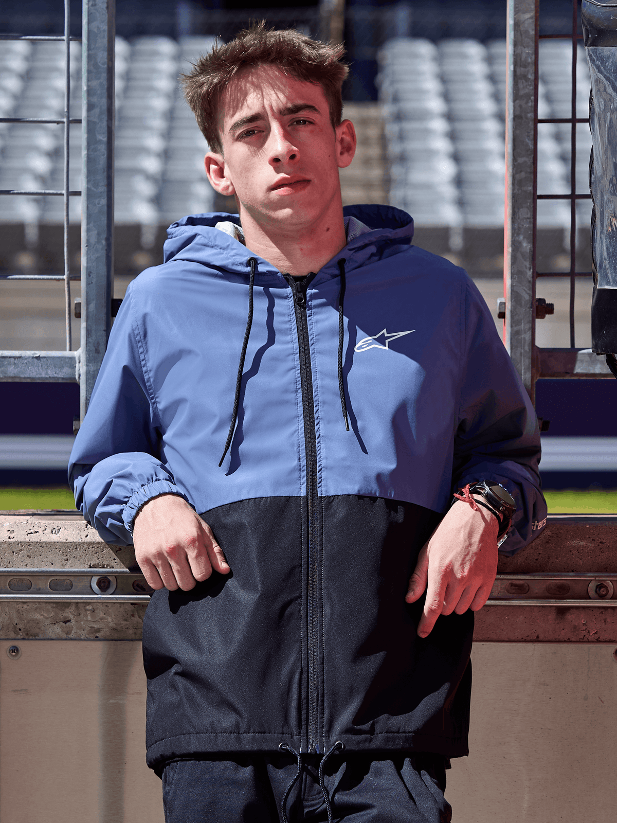 Alpinestars Speeder Windbreaker jas, leigrijs blauw en zwart, windbreaker met capuchon en kleurblok-ontwerp, wit logo op de borst, casual lifestylekleding gedragen op een circuit