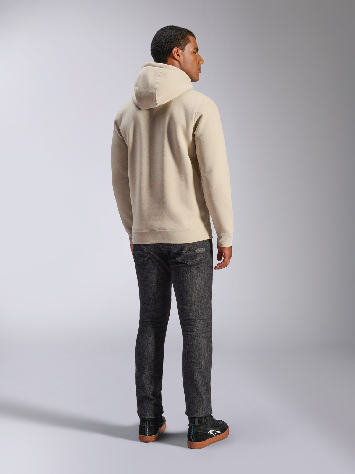 Alpinestars Ageless V3 Hoodie, casual off-white fleece trui met capuchon van de achterkant gezien, voorzien van geribde manchetten en zoom, gedragen met een donkere slim-fit jeans en zwarte Alpinestars sneakers