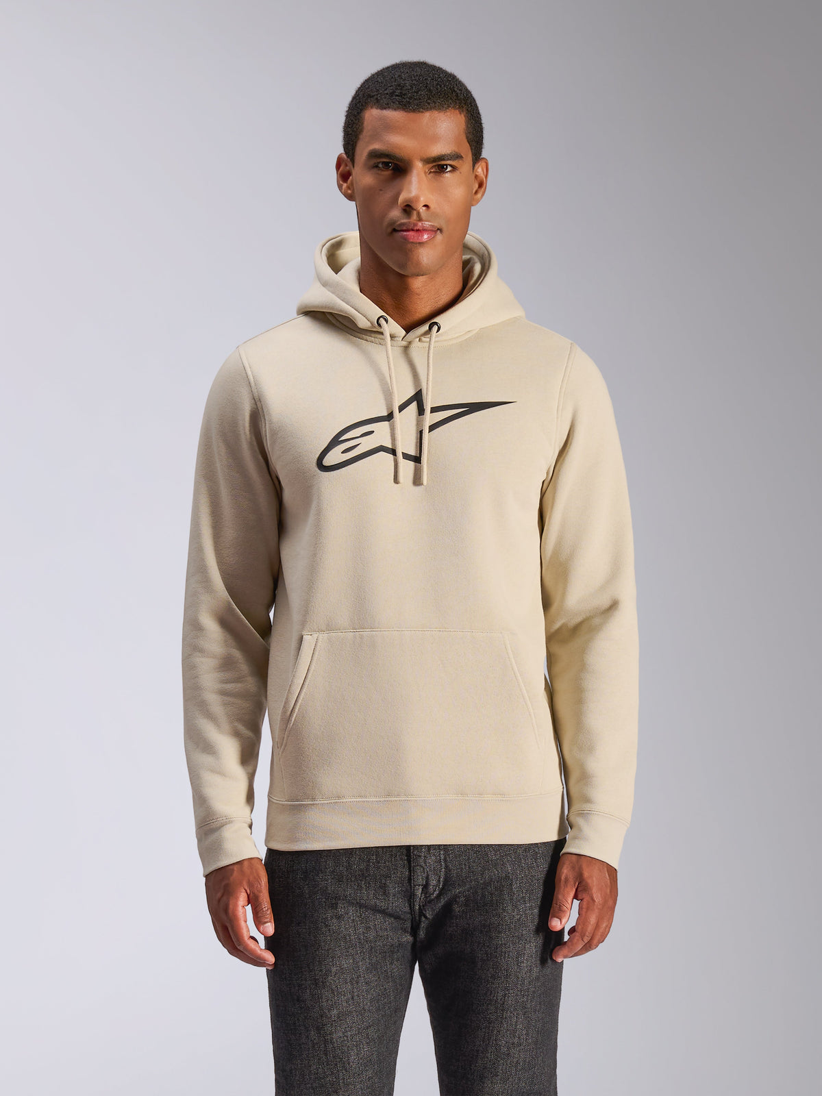 Alpinestars Ageless V3 Hoodie, casual streetwear, gebroken wit met zwart geborduurd logo, pullover met capuchon met trekkoord en kangoeroezak, gedragen door een model met een donkere broek, modern op de motorsport geïnspireerd ontwerp