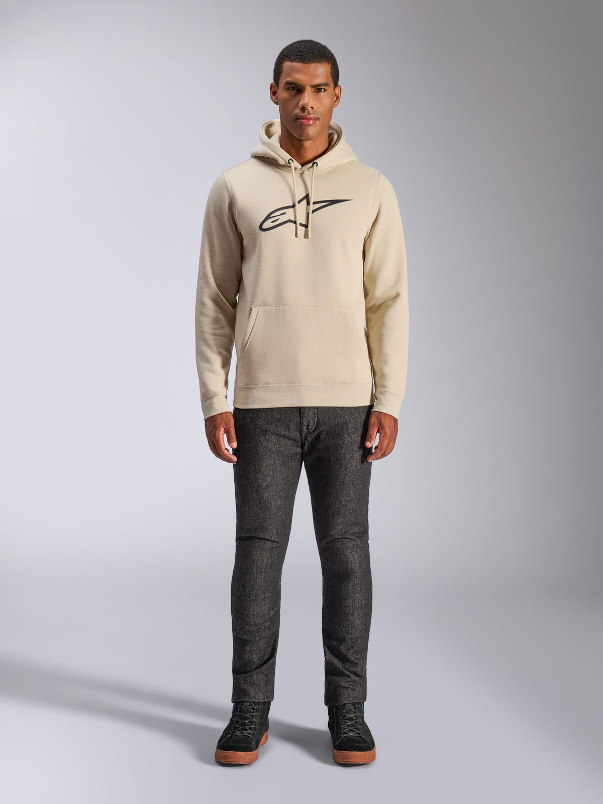 Alpinestars Ageless V3 Hoodie, casual sportkleding, Off White met zwart geborduurd logo, gedragen door een man met een donkergrijze broek en zwarte sneakers, voorzien van een capuchon met trekkoord en kangoeroezak, modern op de motorsport geïnspireerd ontwerp