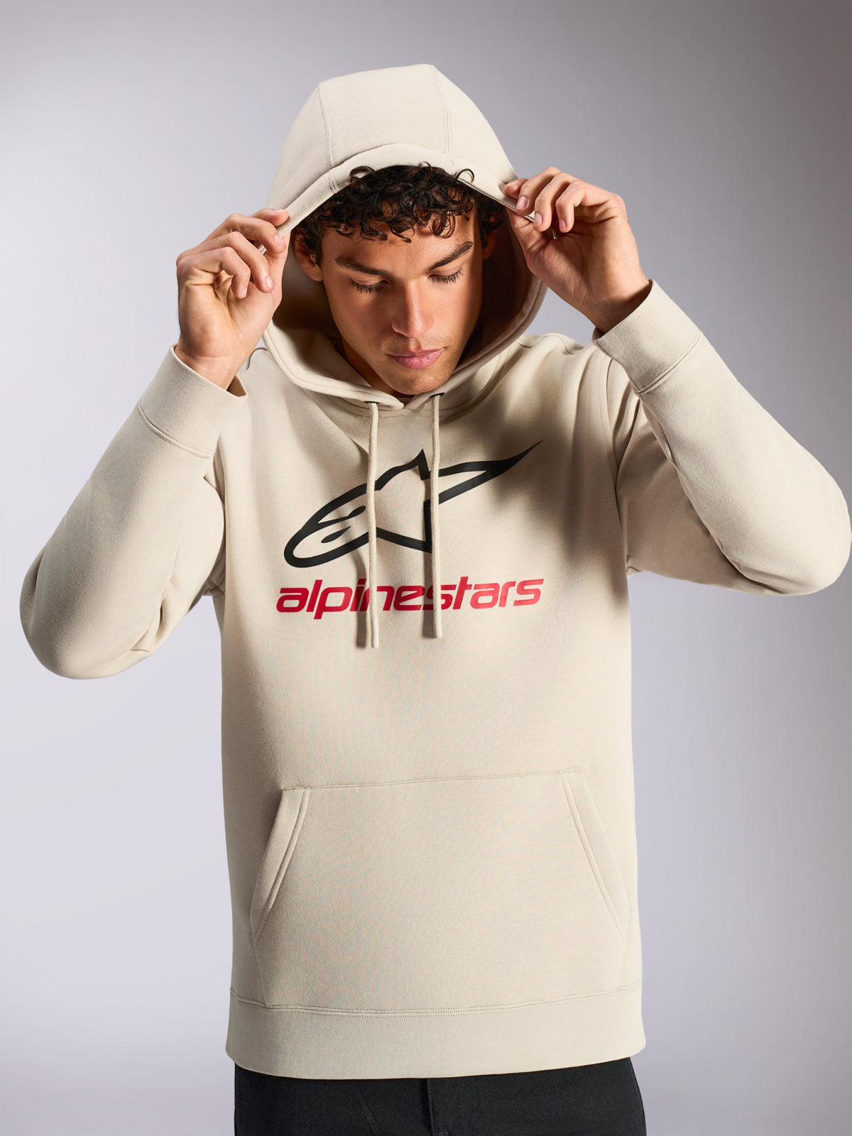 Alpinestars Always V3 Hoodie, mannelijk model draagt een off-white pullover fleece, zwart en rood lineair logo op de voorzijde, capuchon met trekkoord, kangoeroezak, casual motorsport-geïnspireerde streetwear