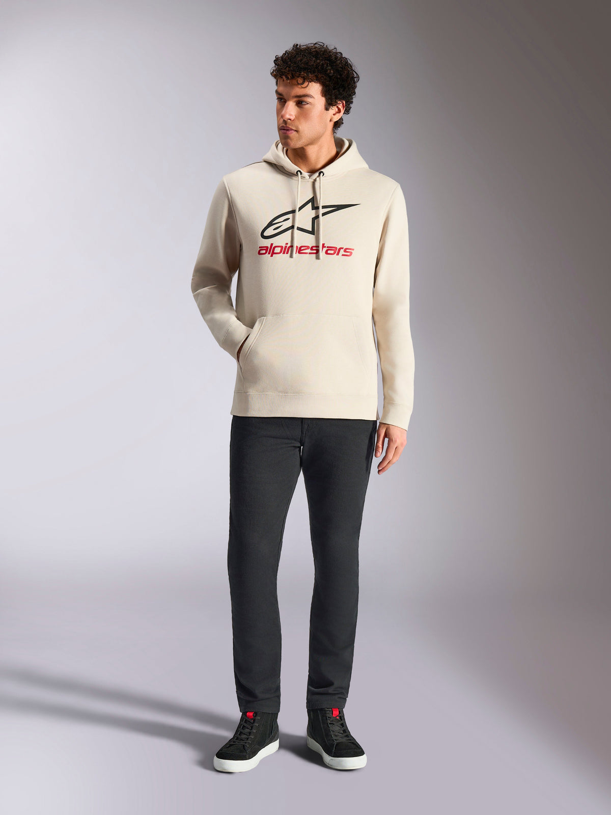 Alpinestars Always V3 Hoodie, casual streetwear, gebroken wit met zwart sterlogo en rode belettering, capuchon met trekkoord, lange mouwen, gedragen door een mannelijk model met een zwarte broek en zwarte sneakers