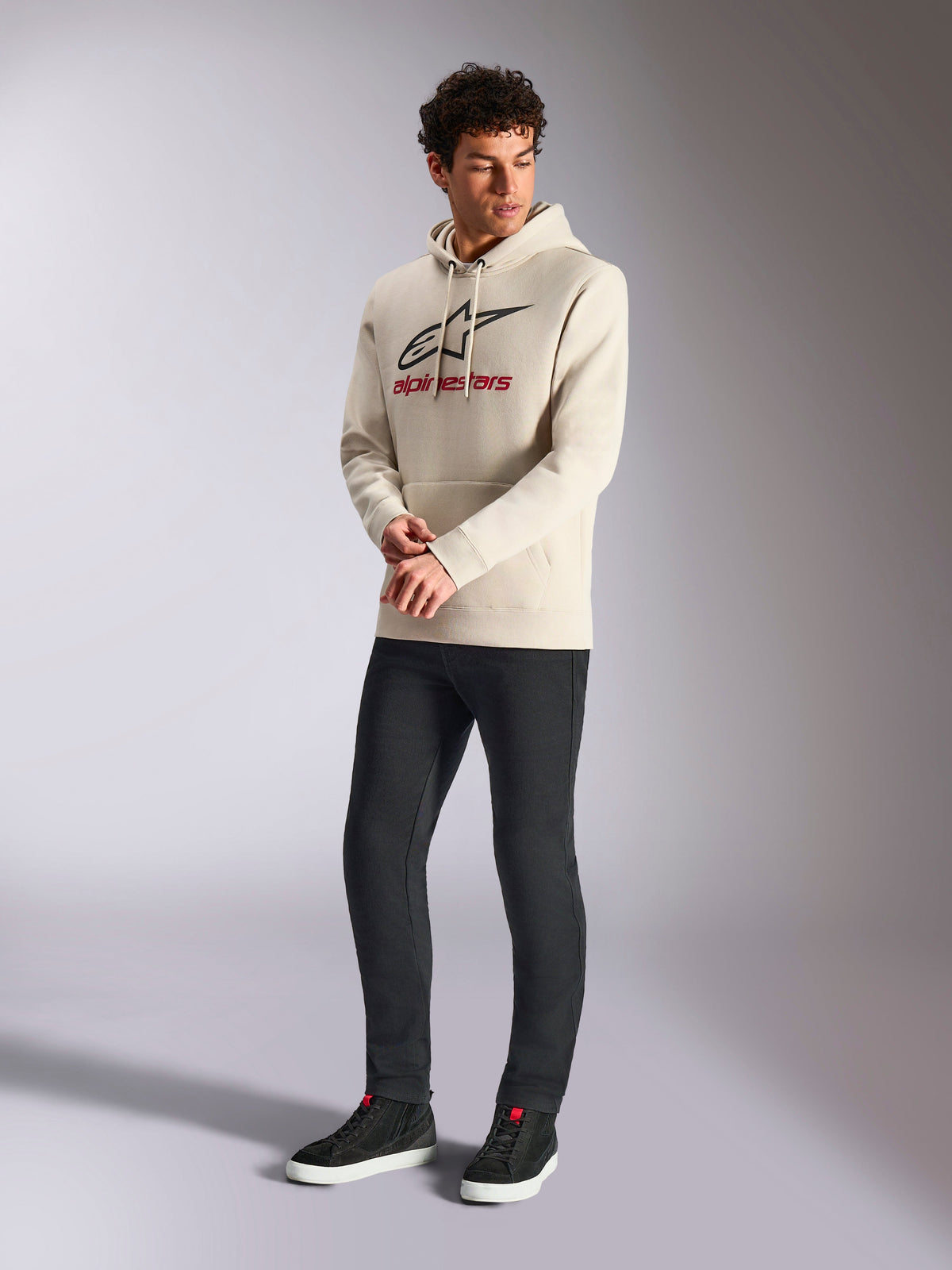 Alpinestars Always V3 Hoodie, off-white pullover fleece hoodie, lineair logo aan de voorkant met zwarte ster en rode merknaam, gedragen door een mannelijk model met zwarte broek en zwarte sneakers, casual motorsport streetwear