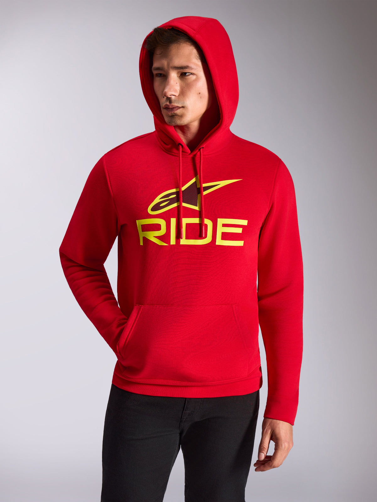 Alpinestars Ride V3 Hoodie, male model wearing red hooded pullover fleece, hi-vis yellow RIDE logo and Alpinestars star on chest, casual motorsport streetwear, front kangaroo pocket, worn with black pants.