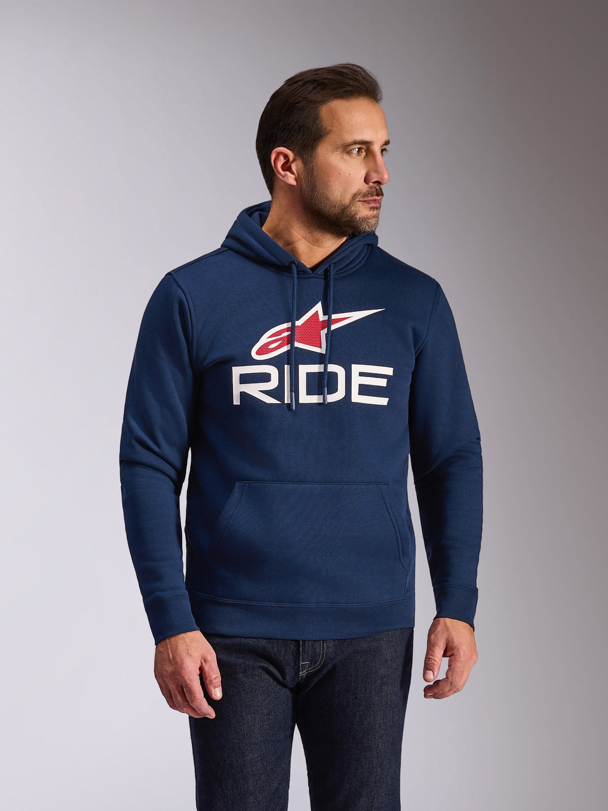 Alpinestars Ride V3 Hoodie, casual marineblauwe pullover fleece, voorzien van een wit RIDE-logo en rood Alpinestars ster-symbool, capuchon met jersey voering en trekkoorden, kangoeroezak aan de voorkant, moderne door motorsport geïnspireerde streetwear, man draagt de hoodie