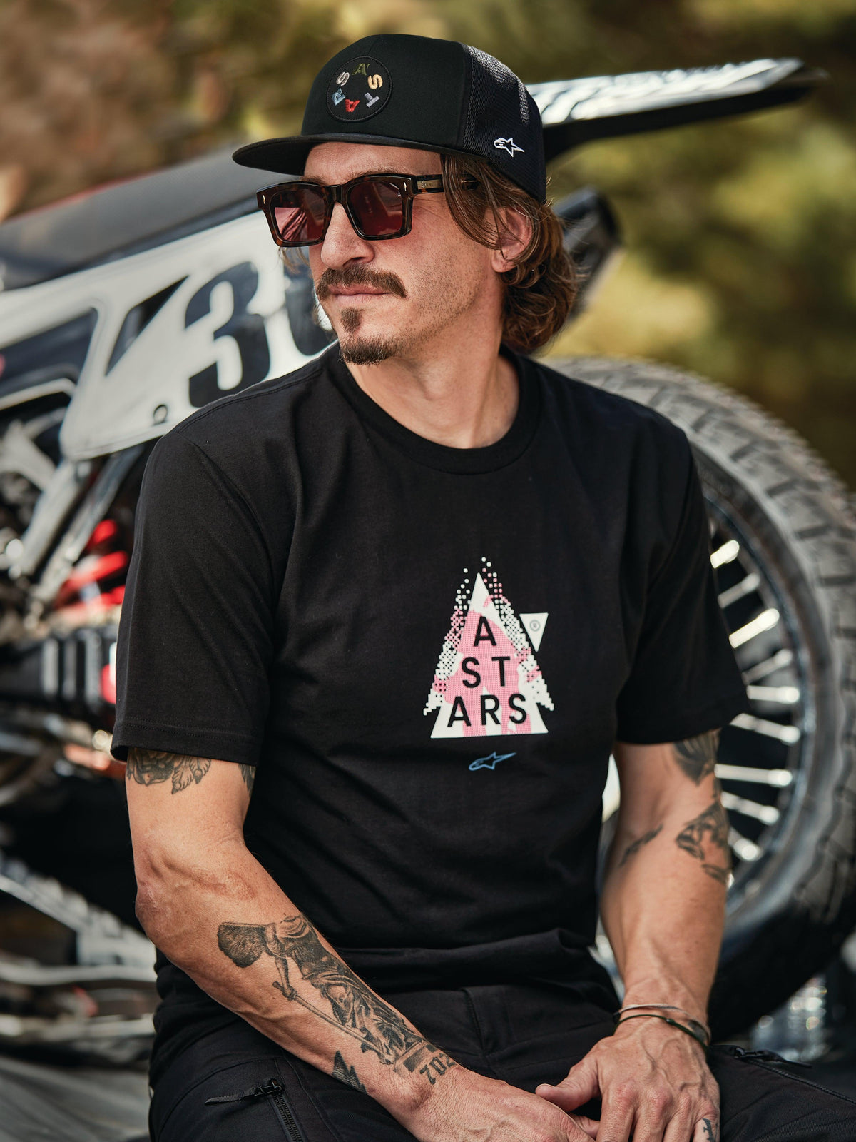 Alpinestars Gambit Trucker pet in zwart met ronde logopatch, gedragen met Alpinestars Soloist SS Tee in zwart, lifestyle-foto van een man met zonnebril en tatoeages zittend voor een motorcrossmotor