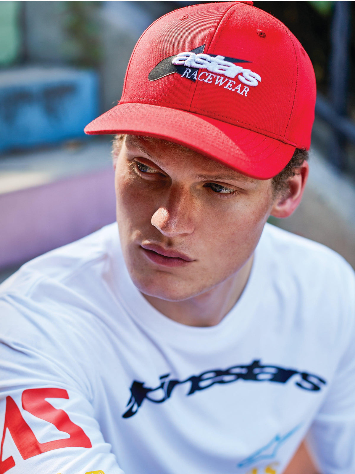 Man die een Alpinestars Profile-pet draagt, rode snapback met gebogen klep en wit en zwart 'astars RACEWEAR' borduurwerk op de voorkant, gedragen met een wit Alpinestars shirt met lange mouwen en graphic, casual motorsport lifestyle-look.