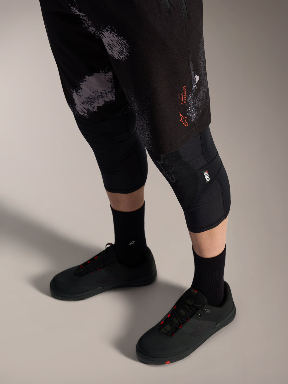 Alpinestars A-Dura Ride MTB-short in Black Camo met oranje logo, gedragen met kniebeschermers en fietsschoenen.