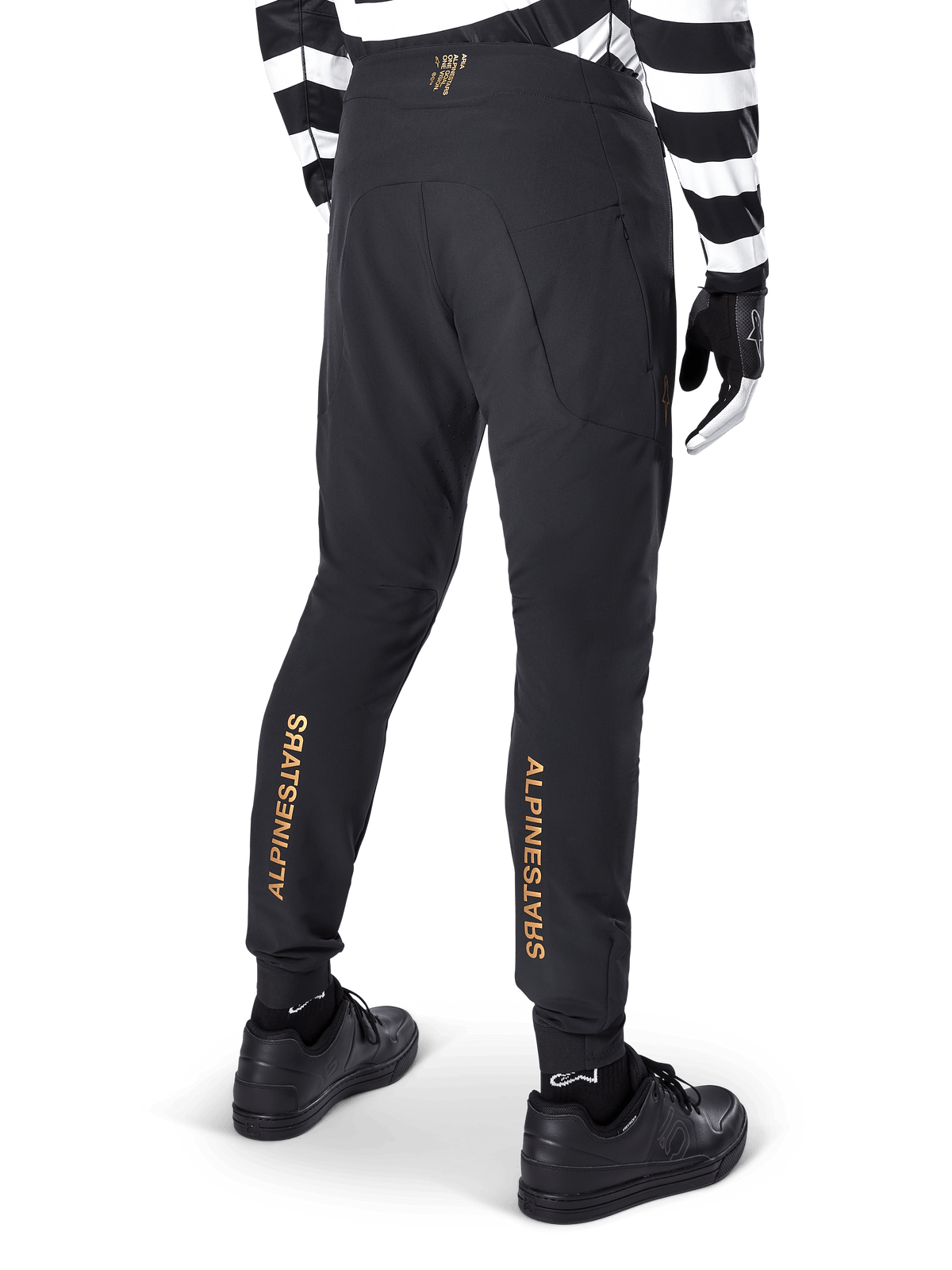 Alpinestars A-Aria Elite broek, zwarte MTB-broek, achteraanzicht, met verticale gouden Alpinestars-branding op de achterkant van de broekspijpen, gedragen met een zwart-wit gestreept shirt en zwarte schoenen, technische fietskleding voor mountainbiken