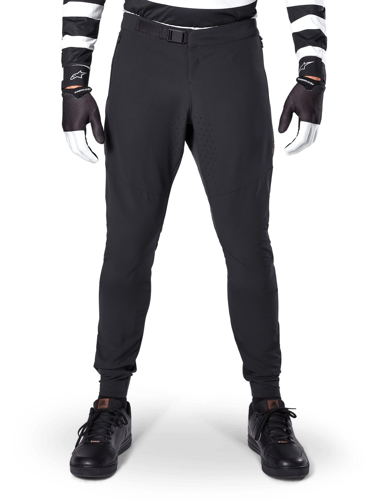 Alpinestars A-Aria Elite broek, mountainbikebroek, zwart, vooraanzicht met laserperforaties en magnetische gespsluiting, gedragen met een zwart-wit gestreepte jersey en zwarte handschoenen, hoogwaardige fietskleding voor trailrijden