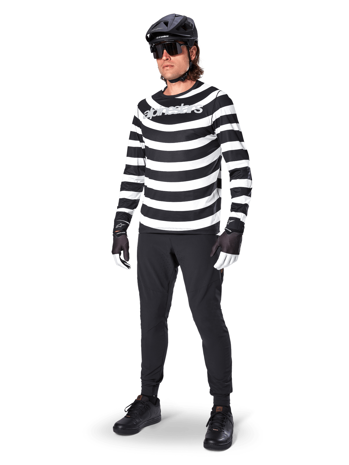 Alpinestars A-Aria Elite broek, zwarte MTB-broek, gedragen door een rijder met een zwart-wit gestreept shirt met lange mouwen, zwarte helm, zonnebril en zwart-witte handschoenen, performancekleding voor off-road fietsen