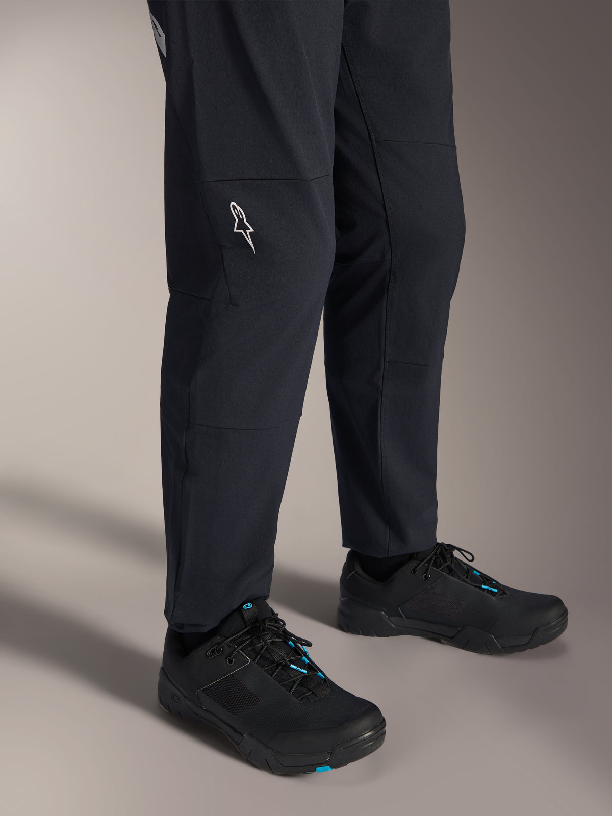 Alpinestars A-Dura Elite Pants, zwarte mountainbikebroek, aanzicht van het onderbeen met een wit Alpinestars-logo op de knie, gedragen met zwart-blauwe fietsschoenen, duurzame constructie voor MTB-rijden
