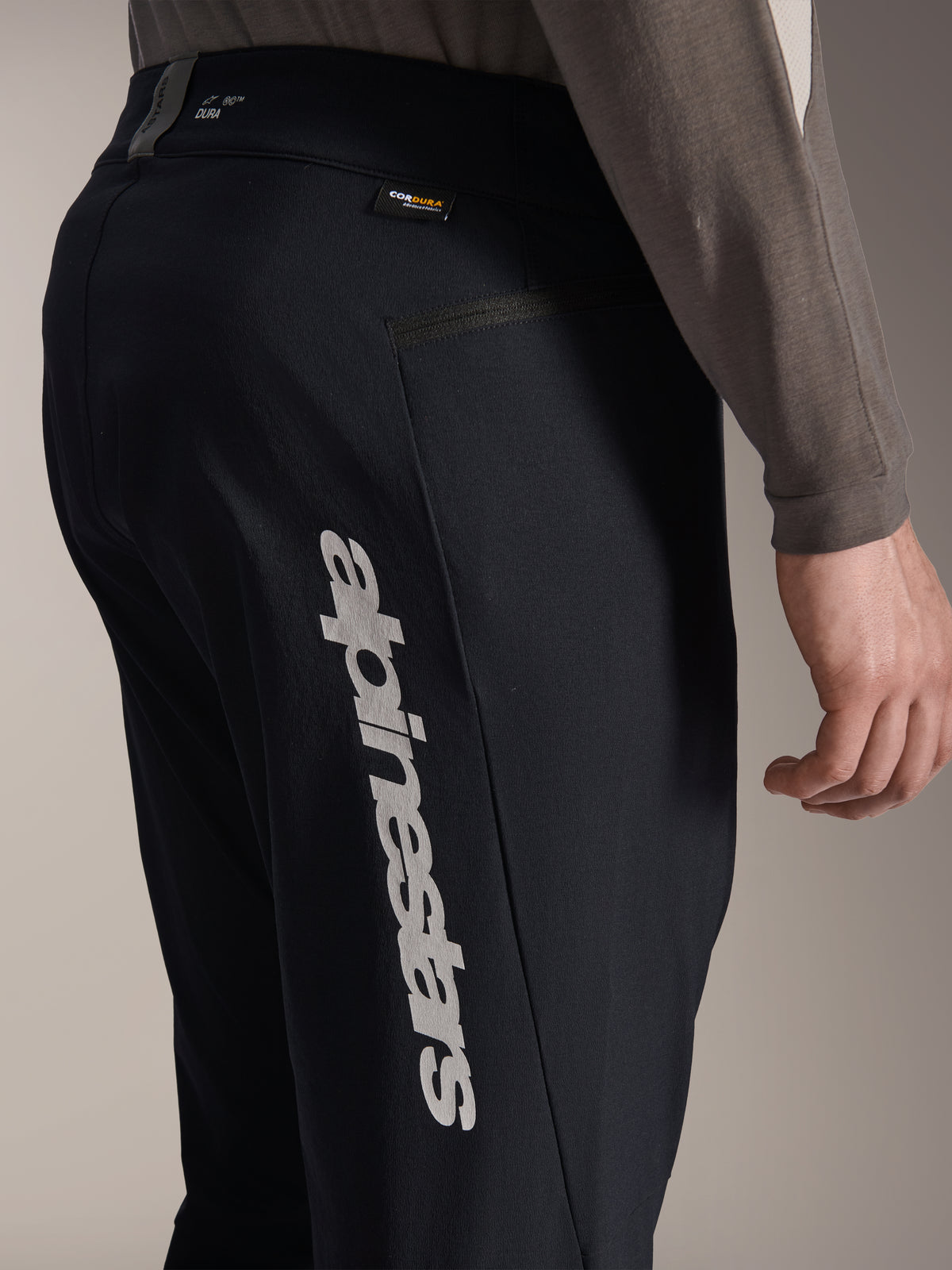 Alpinestars A-Dura Elite broek, zwarte mountainbikebroek, zijaanzicht met groot verticaal Alpinestars-logo op de broekspijp, Cordura-label en zijzak met rits, technische fietskleding voor off-road rijden